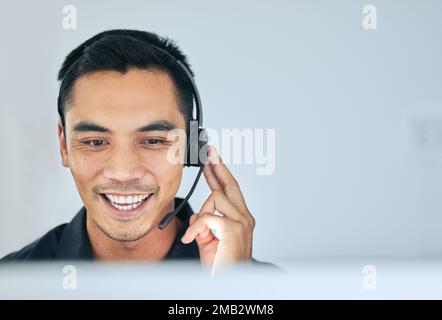 Jeden Anruf mit einem Lächeln entgegennehmen. Ein attraktiver junger Verkäufer, der allein in seinem Büro sitzt und ein Headset trägt, während er seinen Computer benutzt. Stockfoto