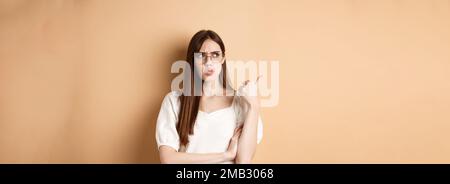 Verwirrt und verdächtig junge Frau in Brille runzelte die Stirn, zeigt und schaut beiseite mit unzufrieden Gesichtsausdruck, stehend auf beigem Hintergrund Stockfoto