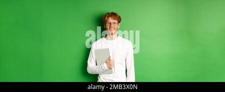 Hübscher junger Mann mit roten Haaren, Brille und Langarm-T-Shirt, Laptop auf grünem Hintergrund haltend Stockfoto