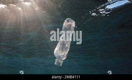 Plastikverschmutzung, weggeworfene Plastikflasche, die unter der Wasseroberfläche in Sonnenstrahlen am Sonnenuntergang driftet. Plastikflasche in blauem Wasser. Plastikmüll Stockfoto