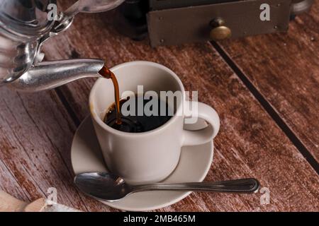 Metallvase, die Kaffee in eine Keramiktasse auf einem Holztisch gießt Stockfoto