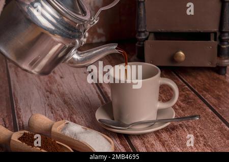 Metallvase, die Kaffee in eine Keramiktasse auf einem Holztisch gießt Stockfoto