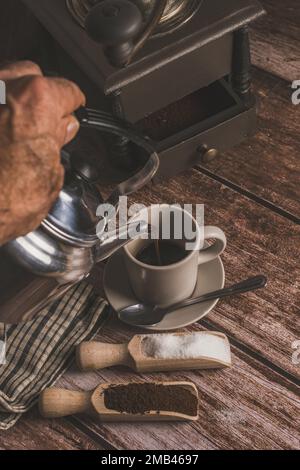 Metallvase, die Kaffee in eine Keramiktasse auf einem Holztisch gießt Stockfoto