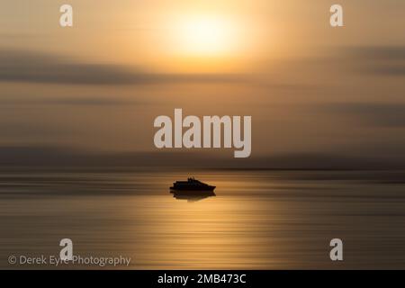 Ein Boot fährt durch die Reflexion der untergehenden Sonne. Absichtliche Wegunschärfe erzeugt einen abstrakten Effekt auf das Meer und den Himmel. Stockfoto