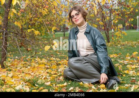 Eine Frau in Lederbekleidung sitzt auf dem Boden inmitten gelber Blätter in einem Herbstpark Stockfoto