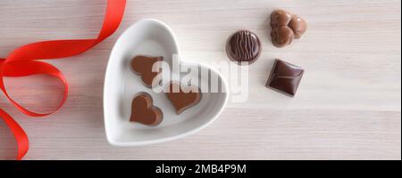 Hintergrund mit einer Auswahl an Schokoladenbonbons in einem herzförmigen weißen Keramikbehälter auf einem Holztisch und einer roten Schleife. Draufsicht. Stockfoto