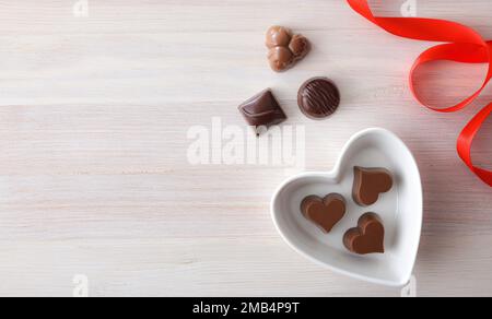 Hintergrund mit einer Auswahl an Schokoladenbonbons in einem herzförmigen weißen Keramikbehälter auf einem Holztisch und einer roten Schleife. Draufsicht. Stockfoto