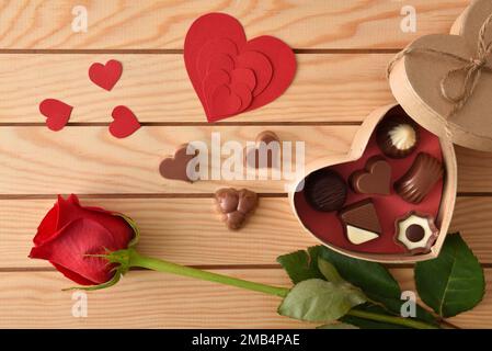 Schachtel mit einer Auswahl an Pralinen und roten herzförmigen Ausschnitten auf einem Tisch mit Holzlatten und einer roten Rose. Draufsicht. Stockfoto