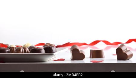 Auswahl an Pralinen, serviert auf einem Teller auf einem weißen Tisch mit verschiedenen herzförmigen Pralinen und einem isolierten weißen Hintergrund. Vorderansicht. Stockfoto