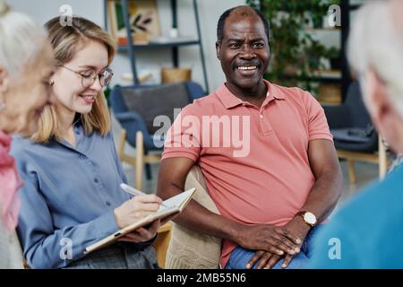 Lächelnder, afroamerikanischer reifer Mann, der Psychotherapiesitzung besucht, er spricht mit einem Spezialisten und anderen Leuten Stockfoto
