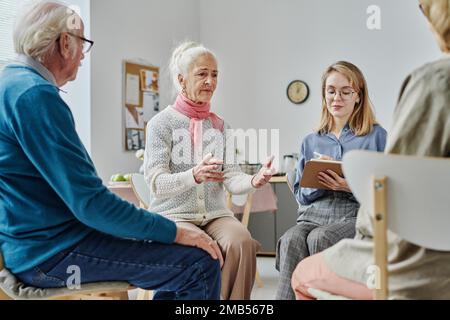 Seniorin besucht Psychologie-Sitzung, sie sitzt auf dem Stuhl und diskutiert ihr Problem mit Psychologen und anderen Leuten Stockfoto
