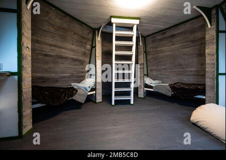 Zwei Einzelbetten mit Bettwäsche in Nischen eines Zimmers. Pyramidenförmiges Bungalow Sommerhaus. Abgewinkelte umgebene Wände, weiße Treppen, grauer horizontaler Arbeitsplatz Stockfoto