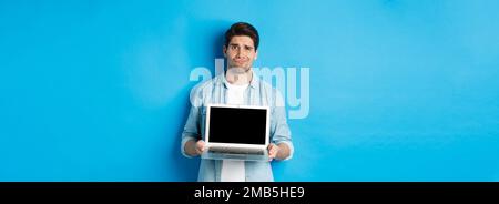 Skeptisch und unzufrieden bärtigen Kerl zeigt Laptop-Bildschirm und grimacing, Zweifel, stehend über blauem Hintergrund in legerer Kleidung Stockfoto