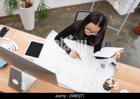 Overhead-Ansicht eines asiatischen Designers, der mit Blueprint und Notizbuch im Büro arbeitet, Stock-Bild Stockfoto