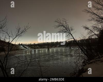 Eine Brücke über den Fluss, die bei Sonnenaufgang von der verschneiten Küste eingefangen wurde Stockfoto