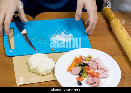 Teller mit Essen, Teig und Küchenutensilien sind auf Holztisch. Das Kind bereitet sich darauf vor, Pizza zu kochen. Master Class in kulinarischen Künsten. Stockfoto