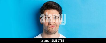 Kopfschuss eines skeptischen Typen, der etwas Unamüsantes, Grimassen ansieht und vor blauem Hintergrund widerstrebend steht Stockfoto