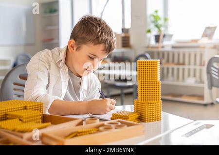 Lächelnder männlicher Junge mit Montessori-Mathematik, goldene Bank, Perlen, alternative Zählentwicklung Stockfoto