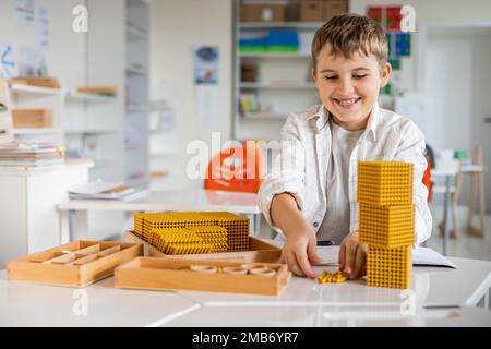 Lächelnder männlicher Junge mit Montessori-Mathematik, goldene Bank, Perlen, alternative Zählentwicklung Stockfoto