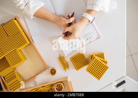 Männliche Kinderhände zählen Mathematik mit goldenen Montessori-Perlen blockieren die Nahaufnahme des Schreibtischs von oben Stockfoto