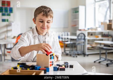 Glücklicher Junge, der Montessori-Stoff-Trinomialwürfel am Klassenschreibtisch zusammenstellt Stockfoto