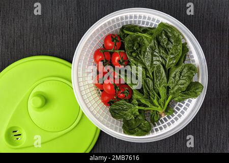 Gewaschene rote Kirschtomaten auf einem Zweig und grüne Spinatblätter in einem Zentrifugensieb auf einem Tisch mit schwarzer Textildecke Stockfoto