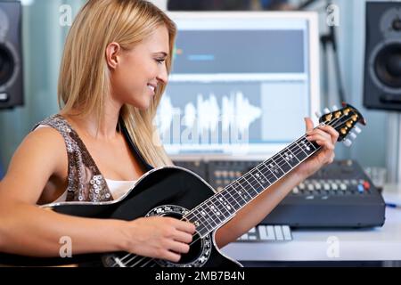 Die Saiten singen lassen. Eine schöne blonde Frau, die in einem Aufnahmestudio Gitarre spielt. Stockfoto