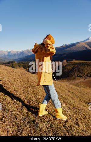 Die Frau geht auf einem Hügel in voller Länge spazieren und sieht die Berge in einem gelben Regenmantel und Jeans im Herbst auf einer schönen Reise bei Sonnenuntergang wandern Stockfoto