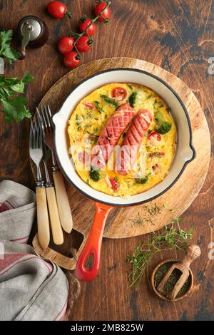 Omelette oder Frittata mit zwei Würstchen, geschmolzenem Käse, Kirschtomaten, Brokkoli und Petersilie auf roter Pfanne oder Eisenpfanne auf altem rustikalem Holzbacken Stockfoto