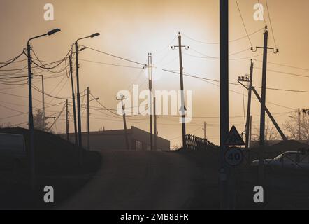 Sonnenuntergang im Dunst über einer Vorstadtstraße mit Strommasten und Straßenlaternen. Stockfoto