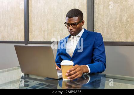 Seriöser afroamerikanischer Angestellter in formellem blauen Anzug und Gläsern mit einer Tasse heißes Getränk zum Mitnehmen am Tisch mit einem Netbook im Büro Stockfoto