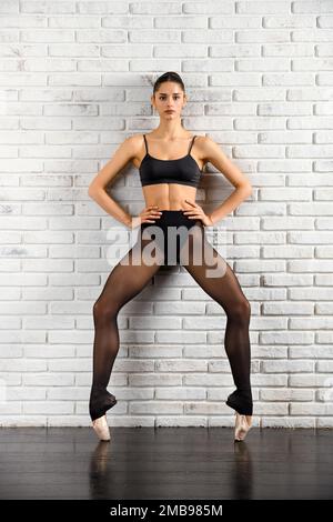 Ganzkörperfit, weiblicher Balletttanz in pointe, der in die Kamera schaut, während er im Studio an einer weißen Backsteinwand steht Stockfoto