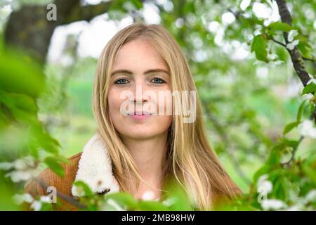 Porträt einer schönen jungen, lächelnden Frau in der Natur mit Frühlingsblüten und zartem grünen Laub auf Ästen, die ihr Gesicht umgeben Stockfoto