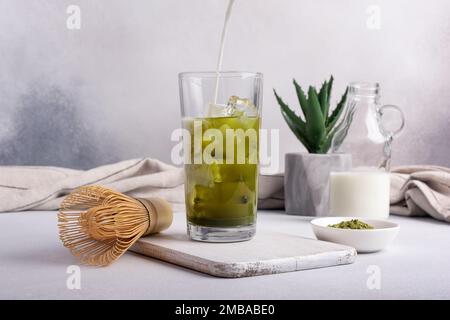 Hausgemachter grüner Matcha-Tee oder Latte mit Eis und Milch oder Sahne in trendy Glas mit Bambusschläger auf grauem Betonhintergrund Stockfoto