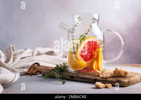 Glas Teekanne oder Wasserkocher für hausgemachten, gesunden Fruchttee mit frischen, reifen Orange-, Grapefruit-, Limonen-, Thymian- und Rohrzucker auf grauem Hintergrund Stockfoto