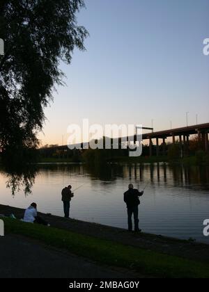 Salford Park, Aston, Birmingham, 2005. Angler, die in der Dämmerung am Aston Reservoir in Salford Park angeln, mit einem erhöhten Abschnitt des Aston Expressway, der Teil des Gravelly Hill Interchange oder der Spaghetti Junction ist und sich am Abendhimmel abspielt. Aston Reservoir ist auch bekannt als Salford Lake, Salford Park Pool oder Salford Bridge Reservoir. Es wurde im 19. Jahrhundert gegründet, um Birmingham Trinkwasser zu liefern. Der See wurde später für Bootstouren und zum Angeln genutzt. Es ist vom Salford Park umgeben. Stockfoto