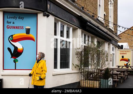 Blythe Hill Tavern, Stanstead Road, Forest Hill, Lewisham, Greater London Authority, 2022. Allgemeine Sicht auf die Ostseite der Blythe Hill Tavern von Südosten, mit einer Frau in einer Covid-19-Schutzmaske neben einem Guinness-Werbetoster im Vordergrund. Stockfoto