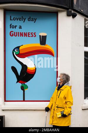 Blythe Hill Tavern, Stanstead Road, Forest Hill, Lewisham, Greater London Authority, 2022. Allgemeine Ansicht einer Frau in einer Covid-19-Schutzmaske, die neben einem Guinness-Werbetoster in der südöstlichen Ecke der Blythe Hill Tavern steht. Stockfoto