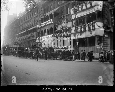 Piccadilly, City of Westminster, Greater London Authority, 1911. Ein Blick nach Westen entlang Piccadilly zeigt Verkehr, Fußgänger und einen Polizisten und Männer auf Leitern, die an der Vorderseite der Gebäude Krönungsdekorationen anbringen. Die Krönungsdekoration in London. An der Vorderseite der Gebäude für die Krönung von König George V. und Königin Mary am 22. Juni 1911 befinden sich Flaggen und Fähnchen. Stockfoto