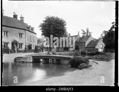 Lower Slaughter, Cotswold, Gloucestershire, 1928. Ein Blick nach Osten entlang des River Eye, wobei ein kleiner Junge auf einer Klapperbrücke sitzt, die auch als flussaufwärts Brücke bekannt ist, mit Häusern dahinter. Stockfoto