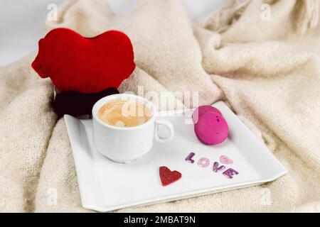 Tablett mit einer Tasse Kaffee im Bett und Macaronkeksen mit Decke, Love-Schriftzug und rotem Herzen, Frühstück zum valentinstag Stockfoto
