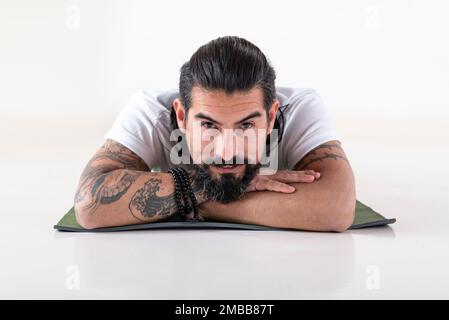 Porträt eines Mannes in weißer Yoga-Kleidung, der auf einer Matte liegt, während er auf einen weißen Hintergrund in die Kamera schaut. Studioaufnahme. Stockfoto