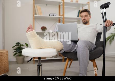 Mann mit gebrochenem Bein, der im Sessel sitzt und einen Laptop benutzt Stockfoto