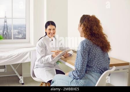 Fröhliche weibliche Ärztin lacht, während sie mit der Frau spricht, während sie ihre medizinische Karte ausfüllt. Stockfoto
