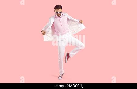 Ein komischer Typ in weißem Anzug, Sonnenbrille und Turnschuhen, die auf pinkfarbenem Studiohintergrund tanzen Stockfoto