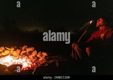 Ein junger Mann bläst Funken in der Nähe eines Lagerfeuers in einer Wüste mit Sternen über dem Kopf. Stockfoto