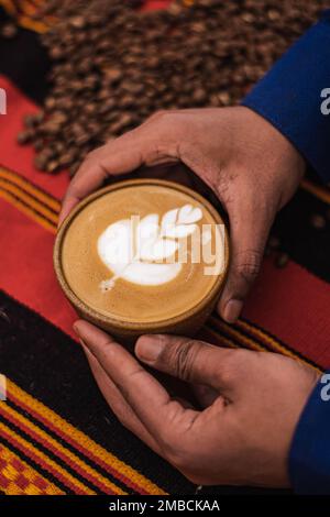 Schwarze Hauthände halten einen Cappuccino in einer Keramiktasse mit Blüten oder Blättern auf dem Schaum in der Hand eines schwarzen Hautmädchens und geröstetem Kaffee Stockfoto