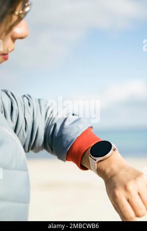 Hüftschuss einer fröhlichen brünetten Frau in Anorak prüft die Fitnessergebnisse auf Smartwatch-Posen am Strand an der Küste. Gesunder Lebensstil Stockfoto