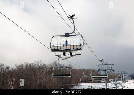 Die Rückansicht eines Snowboarders trotzt den schlechten Bedingungen, wenn er mit einem Skilift auf den Gipfel des Mount Snow fährt und nur sehr wenig Schnee auf dem Berg hat. Stockfoto