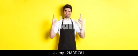 Unzufriedener und skeptischer Kellner mit schwarzer Schürze, der die Finger nach oben zeigt, drückt Abneigung und Gleichgültigkeit aus und steht auf gelbem Hintergrund Stockfoto